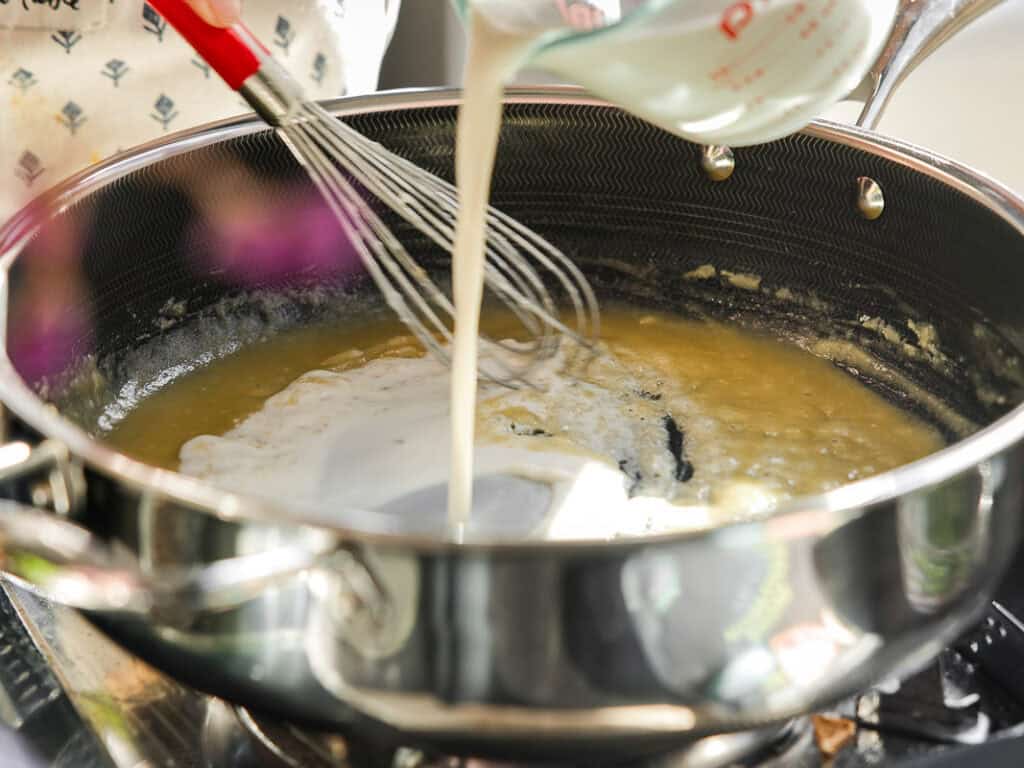 A person is whisking a mixture in a large metal pot on a stovetop while pouring liquid from a measuring cup. The pot contains a light-colored, thick substance. A red whisk is in use, and steam rises from the pot.